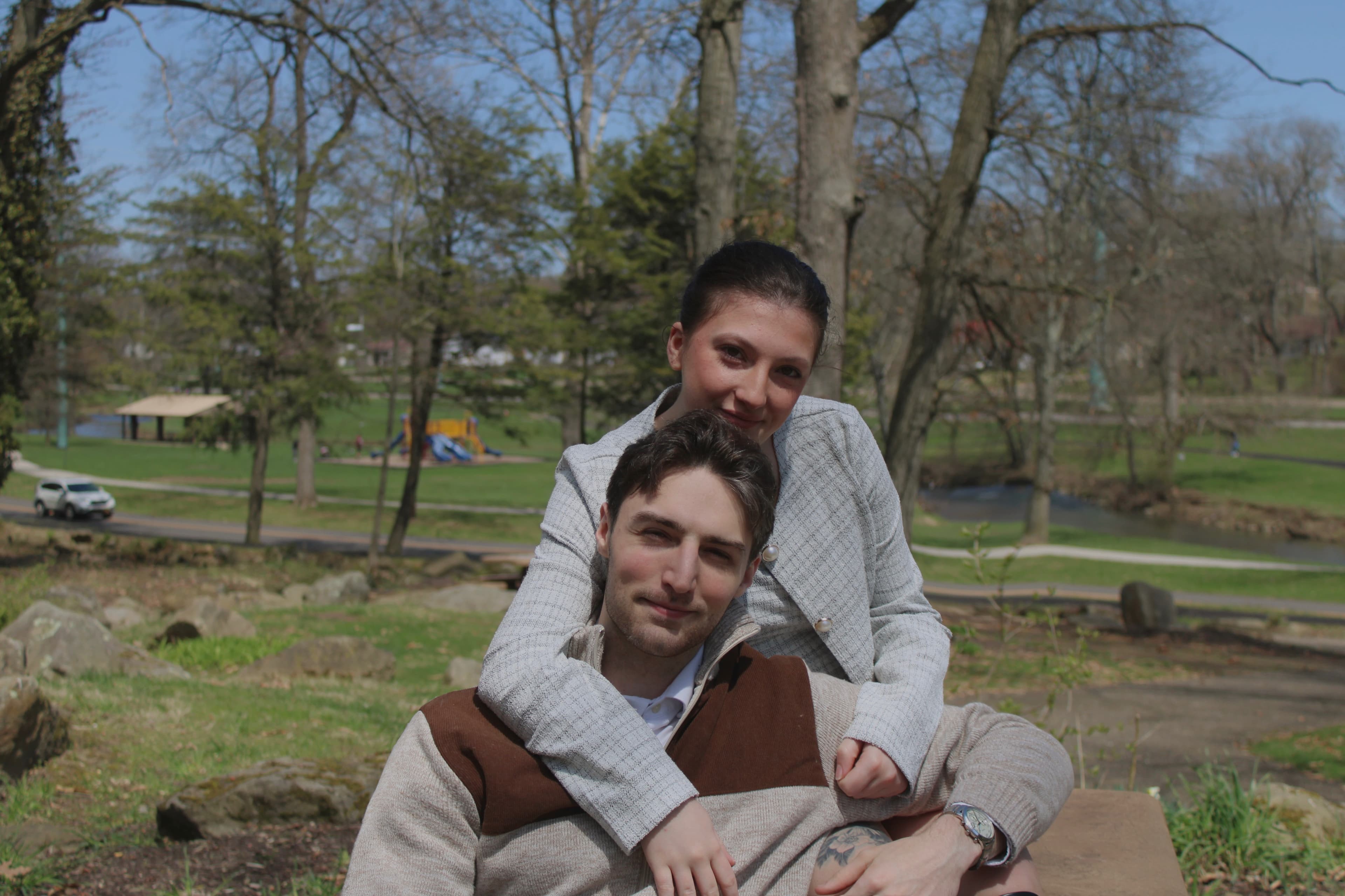 Smiling young couple posing together in a sunny park with trees and a playground.