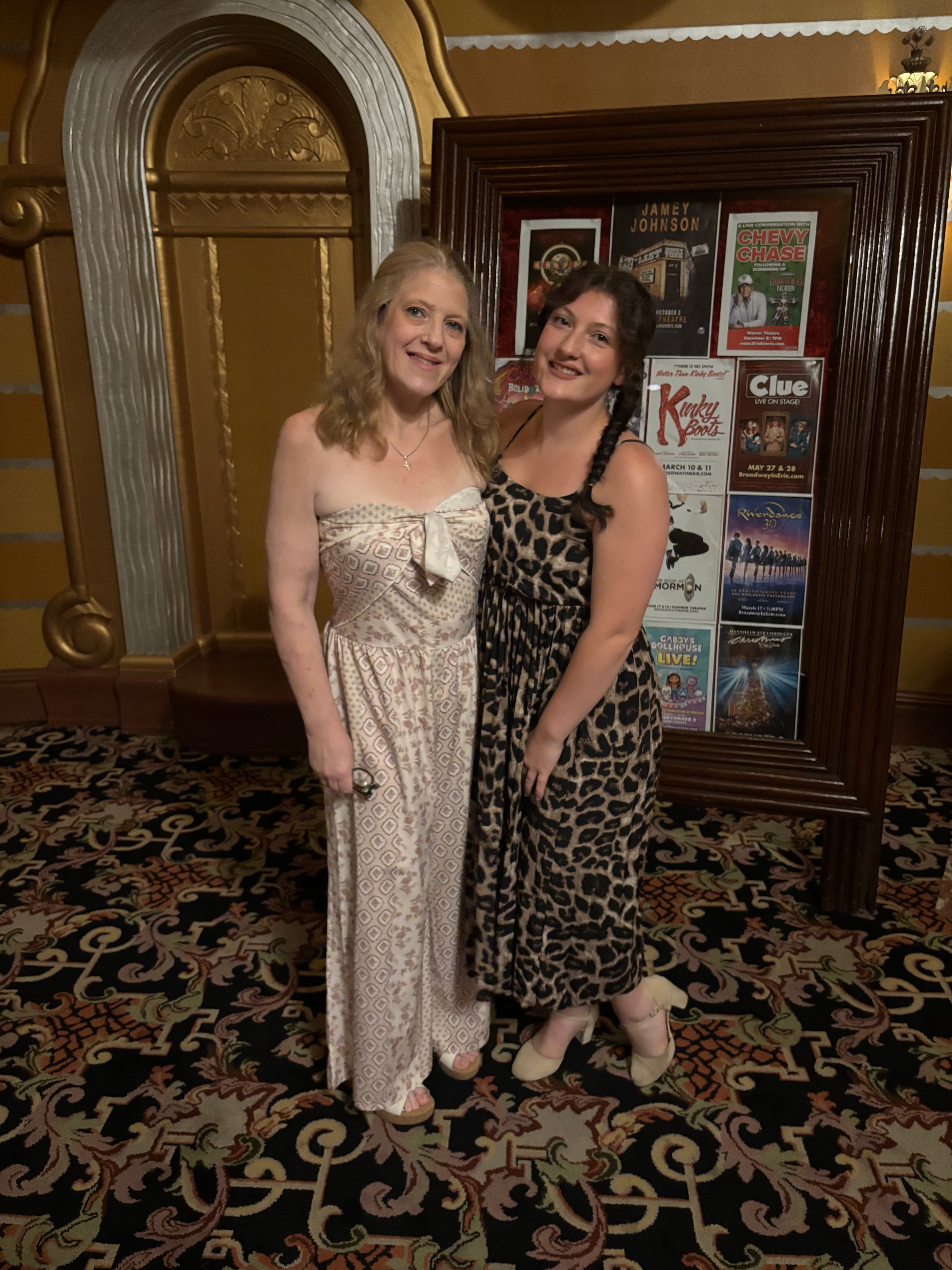 Two women in patterned outfits pose in a theater lobby before a poster display.